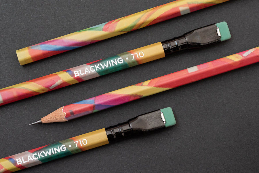 Multicolored Blackwing Volume 710 pencils with erasers, one sharpened, on a dark surface.